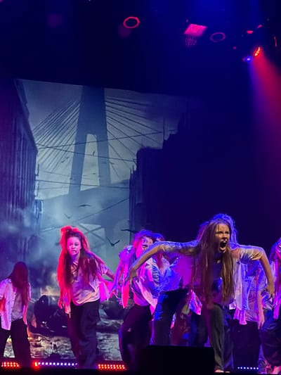 The image depicts a theatrical performance on a stage, featuring multiple individuals in distressed attire against a projected urban backdrop. The backdrop prominently displays a cable-stayed bridge with an A-frame pylon, characteristic of the Ventspils Cable-Stayed Bridge (Venta Bridge) in Ventspils, Latvia. Ruined buildings flank the bridge, and silhouetted birds are visible in the sky.

Approximately eight performers, appearing to be young adults, are present on stage. They are dressed in light-colored, torn shirts with dark pants, suggesting a zombie or post-apocalyptic theme. One central figure, right of the image center, has long hair, an open mouth as if screaming, and arms extended forward. Another figure to the left of the center displays a pale face and intense expression. The performers' poses are dynamic and aggressive, indicating choreographed movement.

The stage is illuminated by dramatic red, blue, and purple stage lighting, with red and blue LED lights lining the front edge. Overhead stage lights are visible in the dark upper portion of the frame, casting colored beams onto the scene.
