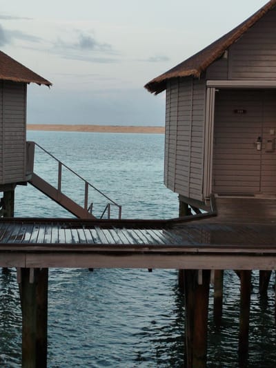 The image depicts two overwater bungalows in the city of Gulhi Falhu, Maldives, situated on stilts above the ocean. The bungalow on the left is partially visible and features a thatched roof and weathered grey siding. The bungalow on the right is more prominent, also with a thatched roof and grey siding. It has a wooden walkway leading to its entrance, which is marked with the number "344" on a small plaque. A set of wooden stairs descends from the left bungalow's deck towards the water. The ocean is a deep blue, with a sandy beach visible in the distance. The sky is overcast with a few wispy clouds. The overall scene is one of serene and tranquil tropical accommodation. There are no people or animals visible in the image.