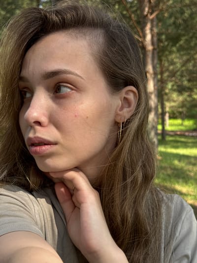 An outdoor, close-up, medium shot shows a young Caucasian woman from the chest up, looking to her left. Her long, brown hair is down and partially obscuring her right ear. She is wearing a beige t-shirt and a gold hoop earring in her left ear. Her left hand is supporting her chin. The background is blurred and appears to be a forest with trees and green grass, consistent with the location of Гольяны, Russia. The lighting suggests it is daytime with sunlight filtering through the trees.