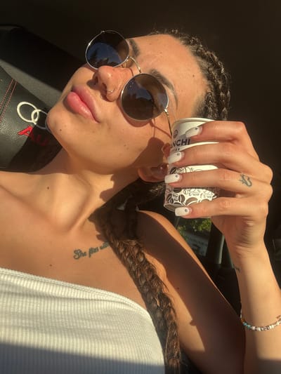 A woman with braided dark hair and a nose ring is in the driver's seat of a car, holding a coffee cup and wearing a white top and sunglasses. She is in Gorna Malina, Bulgaria, and the car interior is visible, including part of an Audi logo on the seat. The lighting suggests it is daytime, possibly late afternoon, with sunlight casting shadows. A tattoo is visible on her chest and another on her arm. She is also wearing a beaded bracelet.