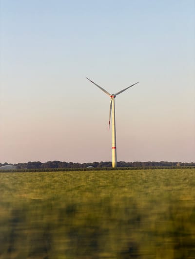 In the city of Geldern, Germany, a single white wind turbine stands prominently in a vast, green field under a clear sky that transitions from pale blue to a soft pink hue. The turbine's blades, tipped with red, are angled upwards and to the sides. A red band encircles the turbine's tower. The field in the foreground is blurred with motion, suggesting movement or a fast shutter speed. In the distance, a line of trees forms a dark horizon against the sky. Sprinklers can be seen in the far distance on the left side of the frame.