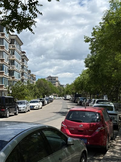 The image shows a street scene in Changping District, China. The view is along a road lined with parked cars on both sides, curving slightly into the distance. The cars are a mix of models and colors, including red, white, and black. Buildings, primarily residential apartments with multiple air conditioning units, line the left side of the road. Trees are present on both sides of the street, creating a partial canopy overhead. The sky is cloudy. No people are directly visible in the frame.