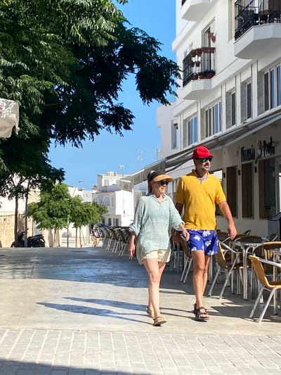 A medium shot captures a couple walking on a sun-drenched, paved pedestrian walkway in Conil de la Frontera, Spain. The walkway is lined with trees on the left and tables and chairs of an outdoor cafe on the right, leading to a white building with multiple stories and balconies. The couple walks side-by-side, the woman on the left and the man on the right. The woman, wearing a light-colored striped long-sleeved shirt, beige shorts, and a sun visor, walks with her left hand at her side. The man, sporting a bright yellow polo shirt, blue floral patterned shorts, a red baseball cap, and sunglasses, walks with his arms at his sides. Both are wearing sandals. They appear to be engaged in casual conversation or simply enjoying a stroll. The sunlight casts long shadows on the ground, indicating it is likely late afternoon. In the background, a scooter is parked near a wall on the left, and a restaurant sign is visible on the building. The sky is clear and blue.