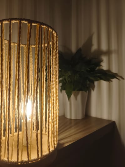 A macrame lampshade with a visible lightbulb is positioned on a wooden surface. Behind the lamp, a green plant in a white pot is situated against a backdrop of light-colored curtains. The scene is dimly lit, with light emanating from the lamp creating shadows on the curtains and the wall. The wooden surface appears to be part of a nightstand or table. The image captures a domestic interior setting.