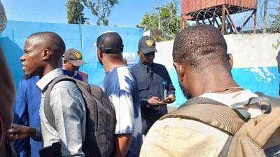 The image captures an outdoor scene in Kabinzi, Rwanda, featuring five adult males under a clear blue sky. In the foreground on the right, the back of a male subject's head and shoulders are visible, showing short dark hair and a beard; he wears a light-colored t-shirt and a tan backpack, with a silver chain around his neck. To his left, a male subject with short dark hair and a beard is seen in profile, facing left with his mouth slightly open, wearing a grey long-sleeved shirt and a black backpack. A dark rectangular object, possibly a phone, is visible tucked into his left hip area. A male in a blue suit is partially visible behind him. Central to the frame, a male subject is viewed from behind/side, wearing a blue and white tie-dye t-shirt and a black mesh baseball cap. To his right, a male subject in a dark blue/black short-sleeved uniform shirt, with an attached lanyard and ID card, wears a dark baseball cap featuring a yellow emblem. He holds a small, light-brown rectangular object, possibly a wallet or phone, in both hands, appearing to examine it. Another male in a dark cap and a high-visibility vest is partially visible further back on the left. The background features a prominent light blue wall with white patches. Behind this, to the right, a reddish-brown corrugated metal structure, possibly a watchtower, is visible, topped with multiple strands of razor wire. Additional razor wire is present along a white wall segment to the left of the corrugated structure. Trees are visible above the background structures.