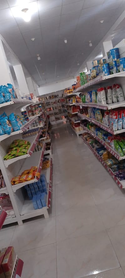 This image depicts the interior of a well-stocked supermarket in Uselu, Nigeria. The scene is dominated by shelves filled with a wide variety of products, suggesting a typical shopping environment. The lighting appears to be artificial, emanating from ceiling fixtures, creating a bright and even illumination throughout the store.
The shelves are organized and neatly stacked with packaged goods, including what appear to be detergents or cleaning supplies in blue boxes on the lower shelves. Higher shelves display bags of snacks, food items, and larger cylindrical containers, possibly representing bulk food products or cleaning detergents. On the far right, more shelves are visible, featuring rows of packaged goods and bottles, suggesting a diverse inventory. The aisle flooring is tiled in a light, neutral color, reflecting the light from above.
There are no people visible in the image, indicating that the photograph may have been taken during a quiet period or before opening hours. The overall impression is one of order and abundance, showcasing a commercial space prepared for customers. The context of Uselu, Nigeria, places the scene within a specific geographical location, likely a busy urban or suburban area where such a store would serve the local community. No specific time of day or weather conditions are discernible from the interior setting.