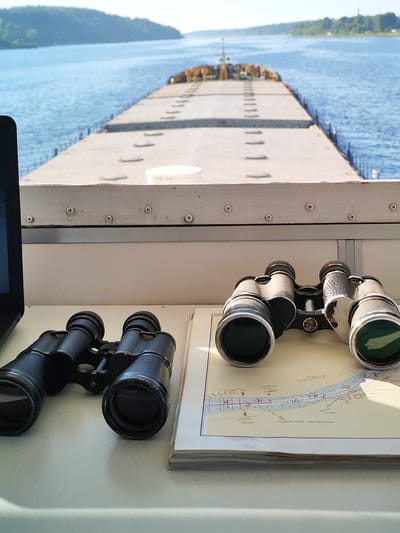 The image depicts a view from inside a cabin, looking out towards a wide, calm body of water. In the foreground, on a light-colored surface, are two pairs of binoculars and a navigation chart. One pair of binoculars is black and positioned to the left. The other pair, silver and black with a metallic sheen, is placed on top of the chart, which appears to be a nautical map with various markings and text. A portion of a laptop is visible on the far left.
Through the window, the expansive water stretches into the distance, bordered by lush green, tree-covered hills on the horizon. The water reflects the bright, clear sky. In the middle ground, a long, flat-topped barge, likely carrying cargo, is visible moving away from the viewer. The barge's deck is a light grey, marked with a series of parallel lines and circular elements. The scene suggests a journey on a waterway, possibly a river or a lake, within the city of Pesochnoe, Russia, given the context provided. The overall atmosphere is serene and focused on navigation and observation.