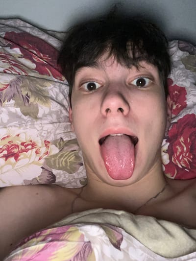 The image depicts an individual in an indoor setting, identified as being in Zainsk, Russia. The subject, an adult with fair skin and short, dark hair, is shown in a close-up, upward-angled portrait, appearing to be lying down. Their head rests on a pillow covered by a floral-patterned case featuring red, pink, green, and yellow elements on a light cream background. A plain light-colored blanket or sheet covers a portion of their upper chest and shoulder. The individual's eyes are wide open, and their mouth is open with a prominent pink tongue extended outwards. A thin, silver-colored chain necklace is visible around the subject's neck. The subject is looking directly into the camera lens. The overall environment suggests a bedroom under low-to-moderate ambient lighting conditions.