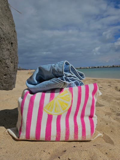 A close-up, low-angle shot captures a beach scene in Yaiza, Spain, focusing on a folded pair of blue denim jeans resting on top of a beach bag. The beach bag features a bold pattern of alternating white and bright pink vertical stripes, with a yellow lemon slice graphic on its front. The items are placed on sandy ground, with the texture of the sand clearly visible. In the background, the turquoise water of the ocean meets a hazy sky filled with scattered clouds. A dark, textured rock formation occupies the left side of the frame, partially obscuring the background. A rocky breakwater is visible in the distance, extending into the sea.