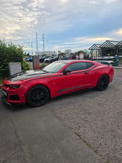 A red Chevrolet Camaro with black racing stripes and a black hood is parked in an outdoor lot. The car is facing left and slightly towards the viewer. In the background, there are several other cars, including a blue one and a white SUV. Several people are visible standing around the lot. Further in the background, there are power lines and what appears to be a fenced-off area with a barrier gate. The sky is cloudy with patches of blue. The scene is likely a parking lot or a gathering area for car enthusiasts. The location is stated to be in the city of Worms, Germany, though this is not visually evident from the image itself.