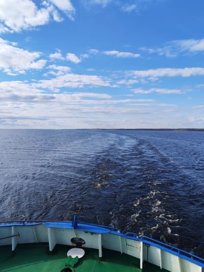 A view from the stern of a boat, presumably on the Volga River near Shchetinskoe, Russia. The sky is blue with scattered cumulus clouds. The water is dark and choppy, with the boat's wake visible behind. The coastline is a faint green band on the horizon. The boat's railing is visible in the foreground, painted with white, green, and blue accents.