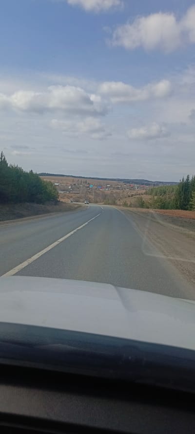 The image depicts a road leading into the city of Zabegalovo, Russia, on a partly cloudy day. The road is paved with a white line separating traffic directions. A vehicle is visible in the distance on the road. The landscape is a mix of trees, fields, and rural buildings with visible red roofs. The photograph is taken from inside a vehicle, with the vehicle's dashboard visible in the foreground.