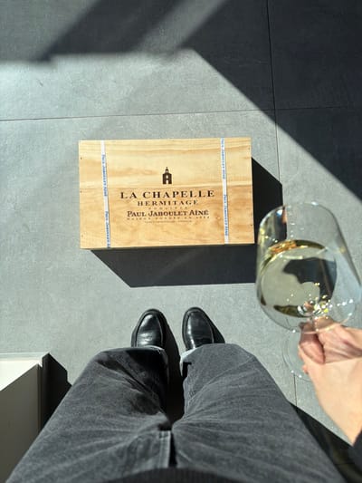 The scene depicts a top-down view of a person's lower body, a wine crate, and a glass of wine on a tiled floor in Marche-lez-Écaussinnes, Belgium. The environment features light gray rectangular floor tiles with visible grout lines. Direct sunlight illuminates the scene from the upper left, casting distinct, angular shadows across the floor and objects, including a large dark shadow in the upper right quadrant. A partial white object, possibly furniture, is visible at the far left edge of the frame.

A light-colored wooden wine crate is positioned horizontally in the upper-middle section. Its visible top surface is printed with a chapel logo and text, including "LA CHAPELLE HERMITAGE DOMAINES PAUL JABOULET AÎNÉ MAISON FONDÉE EN 1834 TAIN L'HERMITAGE - FRANCE." Two vertical blue-and-white straps, bearing the repeated name "PAUL JABOULET AÎNÉ," secure the crate.

Below the crate, the lower half of a person's body is visible. The person wears dark gray or black denim jeans with rolled cuffs and black leather loafer-style shoes on both feet. To the right of the person's right leg, a hand with fair skin tone grasps the stem and bowl of a clear stemmed wine glass, which contains a pale yellow-gold liquid consistent with white wine.