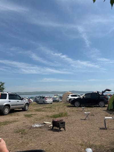 The image depicts a scenic outdoor campsite by a body of water, under a partly cloudy blue sky. The ground is dry and uneven, with patches of sparse grass. Several vehicles, including SUVs and sedans, are parked in the area, suggesting a gathering or event. A portable charcoal grill is set up with embers, indicating recent use for cooking. Three folding chairs are placed around the campsite, some of which appear empty. In the background, a tent and camping equipment are visible, alongside a distant shoreline with rolling hills. The overall atmosphere is relaxed and recreational, with a sense of people enjoying the outdoors. The location appears to be a natural landscape, possibly a park or designated camping area, with the city of Колодезный, Russia in the vicinity.