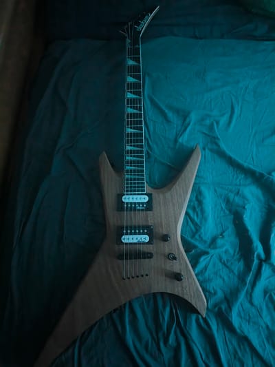 A dark, wooden electric guitar with a pointed headstock rests on a crinkled, teal-colored fabric. The guitar has a sharp, angular body and a dark fretboard with light, triangular inlays. Two black pickups are visible on the body, along with two black control knobs. The headstock features the "Jackson" logo in white script. The lighting is dim and moody, with a blue-green tint, suggesting a low-light environment. The fabric appears to be a bedsheet or a similar soft material, creating folds and shadows around the guitar. There are no people visible in the image. The setting is likely indoors, given the draped fabric and artificial lighting. No time of day or weather cues are discernible.