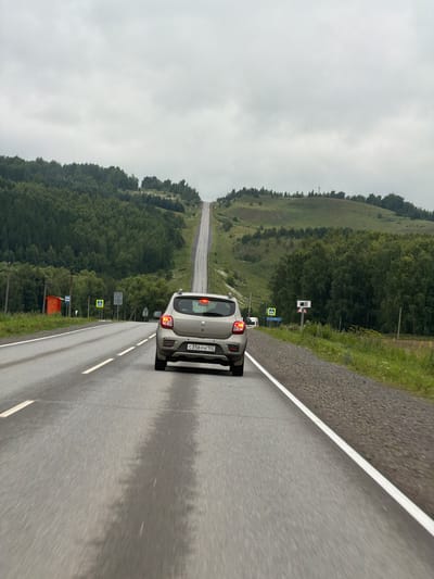 The image shows a car driving on a wet road in a hilly, forested area. The sky is overcast, suggesting it is a cloudy or rainy day. The road has a white line marking the lane. In the distance, the road appears to stretch out into a valley, lined by dense green trees on both sides. There are several road signs visible, including a pedestrian crossing sign. The car, which is light-colored, has its brake lights on. The license plate of the car is visible and reads "C356YH 102". The setting appears to be rural, with a focus on nature and transportation. The overall impression is one of a scenic drive under a grey sky.