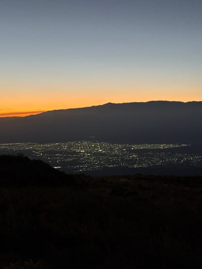 An elevated, wide-angle photograph captures the city of El Portezuelo, Argentina, at either dusk or dawn. The upper portion of the image features a gradient sky, transitioning from light blue at the zenith to a bright orange and yellow hue along the horizon, predominantly on the left. A dark, undulating mountain range forms a distinct silhouette across the mid-horizon, partially obscuring the vibrant sky colors. Below this mountain range, the sprawling city of El Portezuelo is visible, illuminated by a dense network of bright, yellowish-white artificial lights that delineate a clear grid pattern of streets and urban infrastructure. The lower foreground consists of a dark, undifferentiated mass of silhouetted terrain, likely an elevated hillside covered in vegetation, from which the city is observed. No people, discernible objects other than city lights and natural features, specific actions, or interactions are visible.
