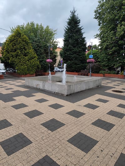 The image depicts a paved public square in Zgierz, Poland, featuring a concrete fountain in the center. The paving stones are arranged in a geometric pattern of tan and dark gray squares. Tall evergreen trees and leafy deciduous trees surround the fountain area, with some buildings visible in the background. A street lamp with a hanging flower basket stands near the fountain. Several cars are parked in the distance, and a bench is situated against a brick border behind the fountain. The sky is overcast.