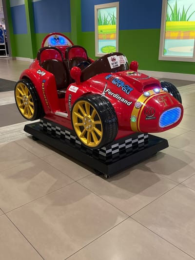 An indoor scene captures a bright red kiddie ride in the shape of a vintage race car. The car features gold-colored wheels, two brown seats, and a blue illuminated grill. The race car is adorned with various decals, including "Golfo," "Rolamte," "Champion," and "Ferdinand." A black and white checkered pattern is visible on the base of the ride, which sits on a beige tiled floor. The background consists of a blue and green wall with two framed artistic depictions of water and grass, possibly representing a park. The overall environment suggests a family-friendly establishment. The location is identified as Maple Ridge, Canada.