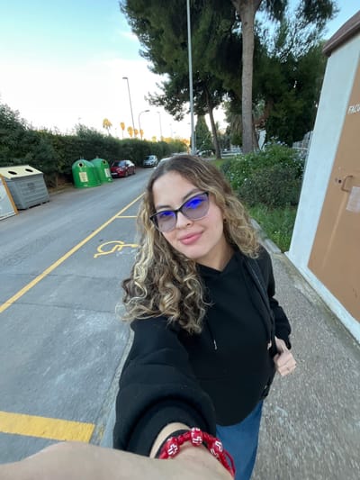 A female, appearing to be in her late 20s or early 30s, with light complexion and long, curly blonde and light brown hair, is captured in a selfie-style photograph. She wears dark-rimmed glasses with reflective blue-tinted lenses, a black hooded sweatshirt, and blue jeans. A red bracelet with white cross-shaped beads adorns her left wrist, and a silver-colored ring is visible on her right ring finger. She is smiling faintly and looking directly into the camera.

The scene is outdoors, during what appears to be late afternoon or early evening, evidenced by the warm, diffused lighting. She stands on a light-colored paved surface, possibly a sidewalk, adjacent to an asphalt road. The road features a double yellow line marking and a yellow painted disabled parking symbol. Along the left side of the road, a grey recycling bin and two green, cylindrical glass recycling bins are present. Further down the road, multiple cars are parked, including a red sedan and a white vehicle. The roadside is lined with dense green hedges and tall pine trees. In the background, numerous palm trees and streetlights are visible against a clear, pale blue sky. To the right of the subject, a section of a light brown panel or building wall is visible, with the partial inscription "FACU" suggesting a "Facultad" (Faculty) building. Beyond this, a grassy area with small blue flowering plants is visible.

This location is identified as the Burjassot-Paterna Campus of the University of Valencia, located in Paterna, Spain.