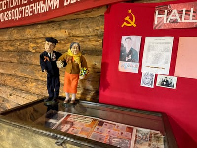The image depicts a museum display case containing a collection of historical documents and photographs. Two life-sized mannequins of an elderly couple, dressed in period clothing and adorned with medals, stand in front of a wooden wall. The male mannequin wears a dark suit and a black cap, while the female mannequin is in a brown jacket and an orange skirt, holding a bouquet of flowers. Behind the mannequins, a red banner with Soviet imagery, including a hammer and sickle, is displayed. Several framed documents and photographs are pinned to the red banner. The display case in the foreground showcases open books with more documents, photographs, and medals. The overall scene appears to be an exhibit dedicated to commemorating individuals and events from Soviet history, possibly in the city of Ust'-Pyzas, Russia, given the context of the provided information.