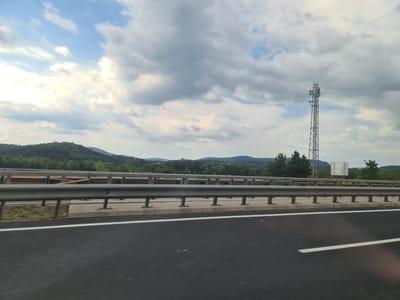 A wide shot shows a cell tower against a cloudy sky, with a lush green hillside and mountains in the background. In the foreground, a dark asphalt highway stretches into the distance, bordered by a metal guardrail and a white line. The scene appears to be on the outskirts of Breitenau, Austria.