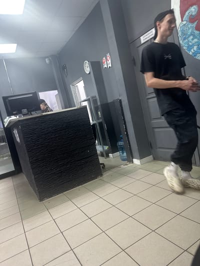 The image shows an indoor scene, likely a business establishment in Donskoy, Russia. In the foreground, a person wearing a black t-shirt, black pants, and white Croc-like shoes is walking towards the right side of the frame. The person is wearing a black cap. To the left, a black reception desk with a textured surface is visible, with a computer monitor and a person behind it. The environment appears to be an office or a service area, with grey walls, tiled flooring, and overhead lighting. A large water cooler bottle is present near the desk, and a clock is mounted on the wall. A sign with Russian text, "СЛУЖЕБНОЕ ПОМЕЩЕНИЕ," which translates to "SERVICE ROOM," is affixed to a door in the background.
