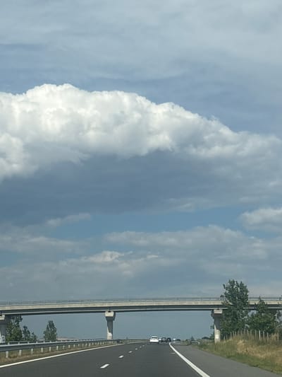 The image depicts a highway scene in Zheleznik, Bulgaria, under a cloudy sky. A concrete overpass, supported by multiple pillars, spans across the highway. Several cars are visible on the road, traveling in the same direction. The highway is paved and marked with white lines. To the right of the road, there is a grassy area with a fence and some trees. The sky is filled with a mixture of white and gray clouds, with patches of blue sky visible. The overall scene is one of typical highway infrastructure in a rural or semi-rural setting.