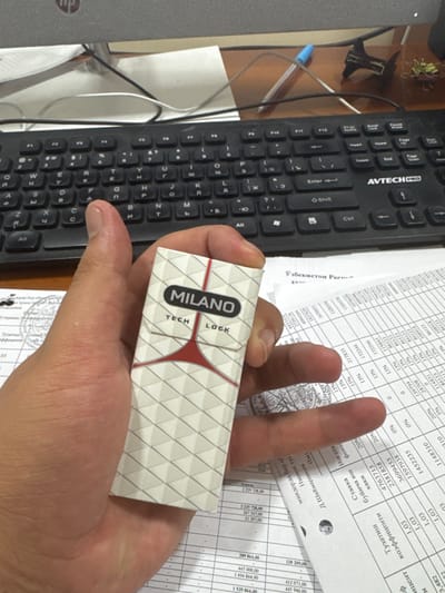 A hand holds a "Milano Tech Lock" package against a cluttered desk. A black keyboard with Cyrillic characters sits to the left. A stack of documents with numerical data and signatures are partially visible in the background. The "Milano Tech Lock" package is white with a grid pattern and a red, triangular design. The documents appear to be financial or official paperwork. The overall setting suggests an office or administrative environment.
