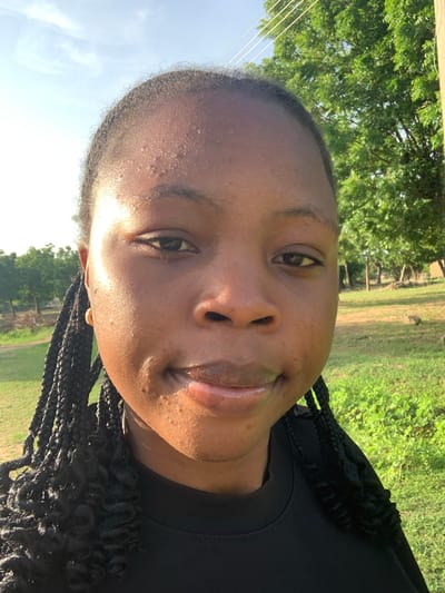 The image shows a close-up, head-and-shoulders shot of a young Black woman with braided hair, wearing a black t-shirt. She is outdoors with a background of trees and green grass, suggesting a rural or suburban environment. The lighting indicates it is daytime, possibly late afternoon, given the angle of the sun. There is no specific information that places the location in Reak, Nigeria. The subject's face is the primary focus, with no discernible actions or interactions. The environment appears natural and uncultivated.
