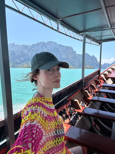 A person wearing a gray baseball cap and a colorful crocheted top is on a longtail boat in Ban Chiao Lan, Thailand. The turquoise water is visible on one side of the boat, with large, green mountains in the background under a clear blue sky. The boat itself is made of wood, with wooden seats and orange life vests visible. Above the person is a metal roof structure.