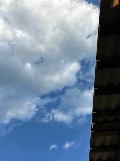 The image captures a view of the sky from under a corrugated metal roof. The roof, with visible wooden rafters, occupies the right side of the frame, casting a shadow over the lower portion of the scene. The sky is a vibrant blue, dotted with various clouds ranging from large, fluffy white formations to smaller, wispy ones. The perspective suggests the photograph was taken looking upwards, possibly from an outdoor shelter or the overhang of a building. There are no people or animals visible in the image. The environment appears to be outdoors, with natural light illuminating the clouds. The location is specified as Парыцкі сельскі Савет, Belarus, indicating a rural or semi-rural setting.