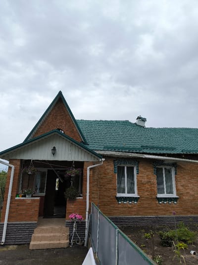 The image depicts the exterior of a residential building, reportedly located in the city of Pysarivka, Ukraine, under an overcast sky. The single-story structure features walls constructed from orange-brown brick with a dark gray, textured foundation course. The roof is composed of green, possibly tiled or shingled, material, with a prominent steep gable over the front entrance. A metal chimney is visible on the main roof section towards the right.

The entrance is accessed via three tan-colored concrete steps leading to a covered porch with a white, gabled overhang. A dark wall-mounted lantern is affixed to the wall above the entrance. Two purple flower hanging baskets are suspended from the porch ceiling, and several small potted plants are arrayed on a ledge to the left of the doorway. The doorway itself appears dark and open. White downspouts are visible along the left side of the porch and below the roofline.

To the right of the porch, two large rectangular windows with white frames are visible on the brick facade. These windows are accented by decorative green and white brickwork forming lintels and sills. Adjacent to the porch steps on the right, a white metal stand supports a large pot containing a pink flowering plant. A low fence with green framing and translucent panels extends diagonally from the lower right foreground towards the house. The ground immediately surrounding the house includes a paved or concrete area near the steps and dark soil with sparse green vegetation, indicating a garden or planted area, to the right of the fence. No individuals are present in the scene.