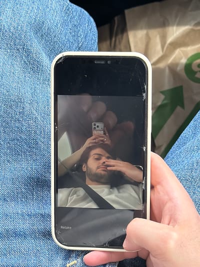 A hand holds a cracked iPhone over denim jeans. The phone displays a selfie photo of a man and a woman. A partially visible green and white bag is in the background. The location is unknown, and there is no evidence suggesting it is Tserovani, Georgia.