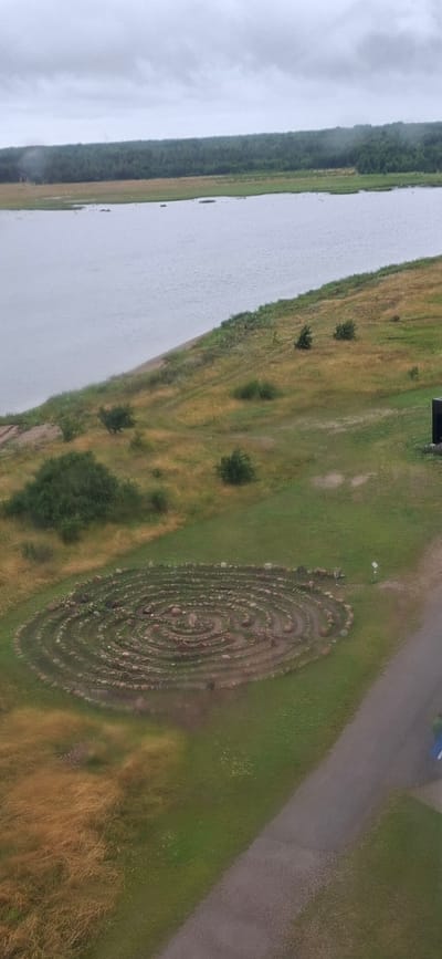 This aerial image, taken from a high vantage point, shows a landscape in Sääre, Estonia. The primary subjects are a body of water, possibly a lake or sea, on the left, and a grassy field with a circular stone labyrinth on the right. The water is a muted grey-blue, suggesting an overcast sky. Along the edge of the water, a dense line of dark green trees creates a natural border.

The field itself is a mix of green grass and drier, golden-brown vegetation, indicating a possible transition of seasons or patchy drought. Several small, dark green bushes are scattered across the field. The most prominent feature in the field is a large, intricately designed stone labyrinth. It is constructed from numerous small to medium-sized stones arranged in a spiraling pattern, creating pathways. The labyrinth is oval-shaped and appears to be a significant man-made structure within the natural setting.

A paved surface, likely a road or path, runs diagonally through the upper right portion of the image, bordering the field. A small white object, possibly a trash bin or marker, is visible near the edge of the labyrinth. The overall impression is one of a serene, rural landscape. The weather appears overcast, given the muted light and the appearance of the water, suggesting it is not midday on a sunny day. There is no visible text in the image. The activity, if any, is not discernable; the image captures a static scene.