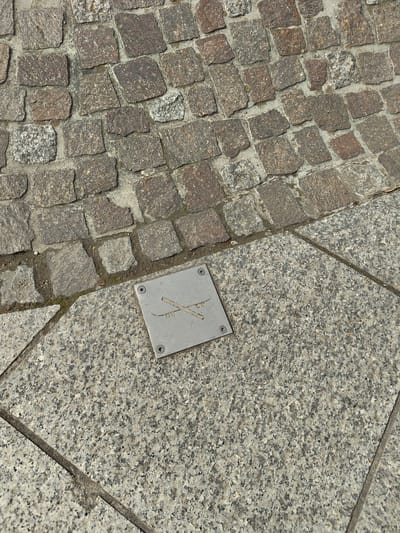 The image displays an overhead, ground-level view of a paved surface in Bertrange, Luxembourg, under daylight. The upper portion consists of irregular, reddish-brown and grey granite cobblestones set in mortar or sand. This cobblestone section diagonally borders a lower area of grey, speckled granite flagstones. A square, brushed metallic plaque is affixed to the granite flagstone surface using four visible screws, one in each corner. The plaque features an etched symbol: two diagonally crossed, elongated lines, resembling ski poles, with two small circular elements and curved lines below them, indicative of goggles. No people or active events are present.