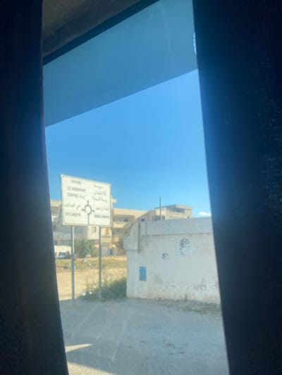 The image depicts an exterior daytime scene captured from inside a vehicle through a slightly tinted window, framed by dark vehicle pillars on the left and right. A clear, bright blue sky dominates the upper half of the view.
In the mid-ground, a white rectangular road sign, mounted on two vertical poles, is prominently positioned. The sign displays directions in both French and Arabic. French text includes "Sousse," "AEROPORT," "CENTRE-VILLE," and "CITE EL BAOUB." Arabic text includes "سوسة," "المطار," "وسط المدينة," "حي البواب," and "MONCEF BEY" beneath a white roundabout symbol. This sign indicates the specific location within or immediately surrounding Monastir, Tunisia, providing directions to its airport, city center, and local neighborhoods, as well as the nearby city of Sousse.
To the right of the sign, a single-story, light-colored building with weathered walls is visible. A rectangular blue marking or patch is present on its left facade, and indistinct markings are faintly visible further right on the wall. The foreground consists of an unpaved ground area with dry, sparse vegetation and scattered debris. In the background, behind the sign and foreground terrain, several multi-story, light-colored buildings are visible under the clear sky. A white vehicle is partially discernible behind the sign on the far left. No individuals or active interactions are visible.