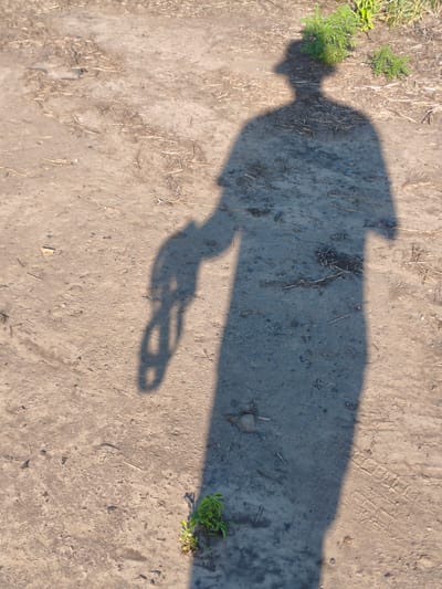 The image shows a shadow of a person cast on a dirt ground. The person appears to be wearing a hat and holding something in their hand, possibly a chain. The ground is dry and dusty, with some small weeds and dry grass scattered around. In the background, there are some green plants growing. The shadow suggests that the sun is low in the sky, indicating either early morning or late afternoon. The scene appears to be outdoors, possibly in a rural or undeveloped area, given the dirt ground and sparse vegetation. There are no visible text elements in the image. The overall impression is one of a solitary figure, captured in a moment of stillness as their shadow stretches across the ground.