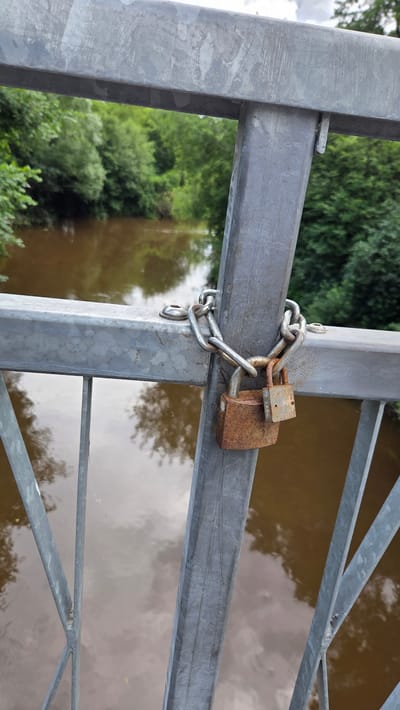 The image shows a close-up view of a metal gate with two rusty padlocks securing it with a chain. The gate appears to be part of a bridge or a fence overlooking a body of water, possibly a river or canal, with murky brown water reflecting the sky and surrounding greenery. The background is filled with lush green trees, suggesting an outdoor natural setting.

The padlocks are made of metal and have a weathered, rusty appearance, indicating they have been exposed to the elements for some time. The chain linking them is also metallic and appears to be of a robust design. The gate itself is constructed from thick metal beams and bars, painted in a dull gray, and shows signs of wear and tear. One of the gate's vertical support beams has a small, flat piece of metal inserted into a gap, possibly a shim or a repair.

The scene is brightly lit, suggesting it was taken during daylight hours. The sky's reflection on the water indicates some cloud cover. There is no visible text in the image. The overall impression is one of security and perhaps a sense of being closed off or restricted, given the locked gate. The location context of Ūlupji, Latvia, places this scene in a rural or semi-rural area of Latvia.
