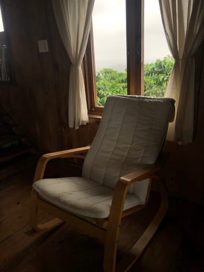 This is a medium shot image of a rocking chair positioned in front of a window. The chair is upholstered in a light-colored fabric and made of light wood. Through the window, a lush green landscape is visible under an overcast sky. The room itself appears to be made of wood, with wooden floors and walls. Light cream-colored curtains frame the window. A light switch is visible on the wall to the left of the window. To the left of the chair, a portion of wooden stairs is visible, partially obscured by shadow. The scene is dimly lit, with the area around the chair and to its left in deep shadow, while the window and the chair itself are more illuminated. The location is stated to be Thôn 4, Vietnam.