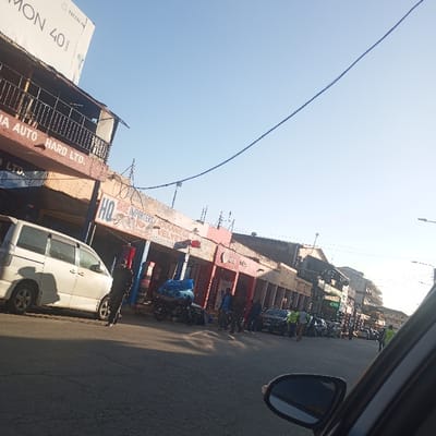 The image displays a bustling street scene in an urban area, viewed from inside a vehicle with its side mirror visible in the bottom right. The scene is brightly lit under a clear, light blue sky, suggesting daytime, likely morning or afternoon.
The street is lined with multi-story buildings, many of which appear to be commercial establishments. A white van is parked on the left side of the road, and several other cars are visible further down the street, some with people gathered around them. Numerous people are present on the pavement and in the road, some moving, others standing. A few individuals are identifiable by their high-visibility vests. Motorcycles are also parked or present near the shops. Overhead wires stretch across the street, indicative of utility infrastructure.
Visible text on signage includes "MON 40" and "Tersas Ph" on a billboard high on a building. Another building prominently displays "A AUTO HARD LTD." and "LTD." A specific shop features "HQ SHOE IMPORTERS KARANTECA VELVET," indicating a shoe retail business.
The image captures typical daily street activity in Sheywe ward, Kenya, showing local commerce and movement of people and vehicles.