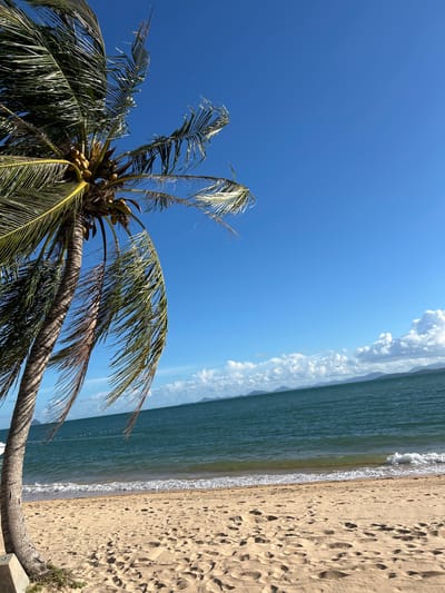 The image depicts a tropical beach scene on a clear day. A palm tree with coconuts stands prominently on the left side of the frame, its fronds blowing in the wind. The sand on the beach is light brown and shows several footprints. Gentle waves are breaking on the shore, and the ocean is a deep blue-green color. In the distance, a hazy shoreline with mountains can be seen under a bright blue sky with scattered white clouds. The location is identified as Ban Khlong Hia, Thailand.