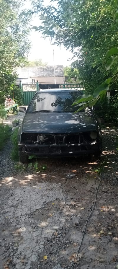 This is a frontal, eye-level shot of a dark-colored car, likely a hatchback, that appears to be abandoned or in a state of disrepair. The car is parked on a gravel and dirt surface, with lush green foliage and trees surrounding it, creating a shaded, overgrown environment. The front of the car is missing its bumper and grill, exposing internal components. The windshield is intact but reflects the surrounding trees and sky, with dappled sunlight illuminating parts of the car's hood.

To the left of the car, a red bicycle is leaning against a green wooden bench. Beyond the car and bicycle, a green corrugated metal fence and a section of a light-colored building with a gray roof are visible. The overall impression is one of neglect and abandonment in a rural or semi-rural outdoor setting. The time of day appears to be daytime, likely late morning or early afternoon, given the bright sunlight filtering through the leaves. The weather seems pleasant and sunny. There is no visible text in the image. The location is specified as Marinovka, Kazakhstan.