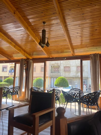 This is a photograph taken inside a café or restaurant, looking out through large windows onto a street scene in El Hajeb, Morocco. The interior features a warm, wooden ceiling with exposed beams and a hanging lantern. In the foreground, wooden chairs with dark upholstery are visible, suggesting a seating area.
Through the windows, we see a street with buildings and trees. One building has a yellowish facade and balconies with laundry hanging. Another building is made of concrete blocks. Several people are on the street, including a woman in a headscarf, another woman, and a person pushing a stroller. Green, globe-shaped bushes are planted outside the windows. The overall impression is of a pleasant, sunlit outdoor seating area connected to a street in a Moroccan town.