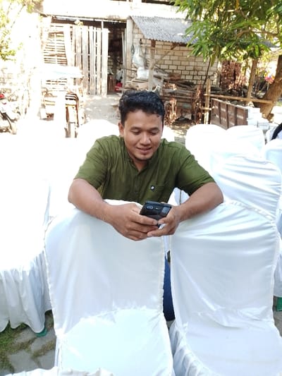 A male subject is depicted in an outdoor setting within the city of Juwana, Indonesia. He is positioned leaning forward, with his forearms resting on the back of a white fabric-covered chair. He has dark, curly hair, a light beard, and is wearing an olive-green short-sleeved collared shirt. His head is tilted downwards, and he is looking at a black smartphone held horizontally with both hands, exhibiting a faint smile. Surrounding him are multiple similar white fabric-covered chairs, indicative of an event setup. In the background, to the left, a light-colored motor tricycle and a partial white scooter are visible near a wooden slatted structure. To the right, a building with light-colored brick or block walls and a corrugated roof is present, surrounded by stacks of various materials, including wood and potentially bricks. A tree with green foliage provides natural shade on the right side of the frame. The ground appears to be a mix of dirt, gravel, and some light-colored paving stones. The scene is illuminated by bright daylight.