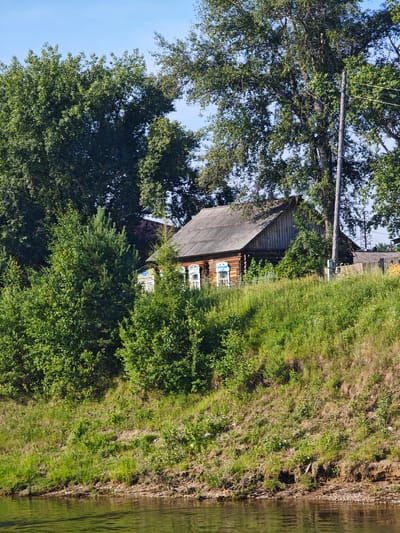 The image depicts a rustic wooden house with decorative window frames situated on a grassy hill overlooking a body of water. Tall trees frame the scene, with a wooden utility pole to the right of the house. The sky is clear and blue. The overall impression is of a tranquil, rural setting, likely in the village of Galanino, Russia, given the architectural style and natural surroundings. There are no people or animals visible in the image.