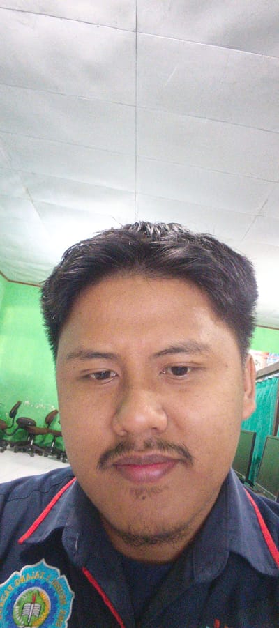 A medium shot selfie features a man with dark hair and a mustache. He is wearing a dark blue collared shirt with red trim and an emblem on the left chest. The man is looking directly at the camera with his eyes open. Behind him, a bright green wall is visible, and further back, several office chairs on wheels are arranged in a row. The ceiling appears to be made of white acoustic tiles. The image quality is grainy, suggesting it was taken in low light conditions. The emblem on the shirt displays text and imagery that appears to be a seal or logo, with a book and a torch prominently featured. The overall setting suggests an indoor environment, likely an office or classroom, possibly in Sukabumi, Indonesia, given the presence of what might be an Indonesian educational or institutional logo.