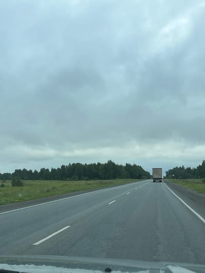 The image captures a view from a vehicle on a paved highway, likely in Kanashevo, Russia, on an overcast day. The highway stretches into the distance, flanked by lush green fields and dense forests on either side. A white truck is visible traveling ahead on the highway. The sky is filled with thick, gray clouds, suggesting a cloudy or potentially rainy day. The road surface appears wet, with reflections visible. The scene is devoid of any visible pedestrians or other vehicles besides the truck.