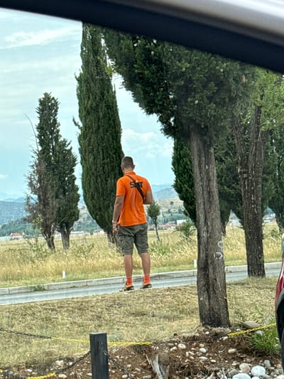 A man wearing an orange polo shirt with "SIXT" printed on the back and khaki shorts stands facing away from the camera, looking towards a landscape of trees and buildings. He is wearing orange socks and brown shoes. He has a black bag slung over his shoulder. The man is standing on a grassy area next to a paved road. In the background, there are several tall, thin trees, some buildings, and hills. The sky is cloudy. The foreground shows a chain-link fence with yellow caution tape. The image appears to have been taken from inside a vehicle, as indicated by the dark frame at the top of the image. The location is identified as Golubovci, Montenegro.