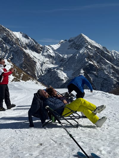 This image is rotated 90 degrees counter-clockwise. It captures a sunny day on a snow-covered mountain, likely a ski resort in Alagna Valsesia, Italy, with towering, rugged, snow-capped peaks forming a dramatic backdrop under a clear blue sky.
Several individuals are present on a groomed ski slope, which shows distinct parallel lines in the snow. On the right side of the frame, three people are seated in black foldable chairs with green accents. The uppermost person is wearing bright yellow ski pants and white ski boots, with "HEAD" visible on one boot. They are positioned with their legs pointing upwards, facing away from the viewer. Below them, two individuals in dark winter jackets are seated side-by-side, looking towards the left and upwards, appearing to admire the view. Partially visible next to them are a pair of skis and a ski pole. Above the seated group, another person in a blue jacket and black pants is partially seen, appearing to stand or move near the chairs.
On the lower left side of the frame, a person wearing a red, white, and black ski jacket and black pants stands, holding a mobile phone. They are also looking towards the left, suggesting they are capturing the mountainous landscape.
The overall scene conveys a sense of relaxation and enjoyment amidst a stunning alpine environment, with people taking a break to appreciate the panoramic views. The bright daylight and clear sky indicate excellent weather conditions for outdoor activities.